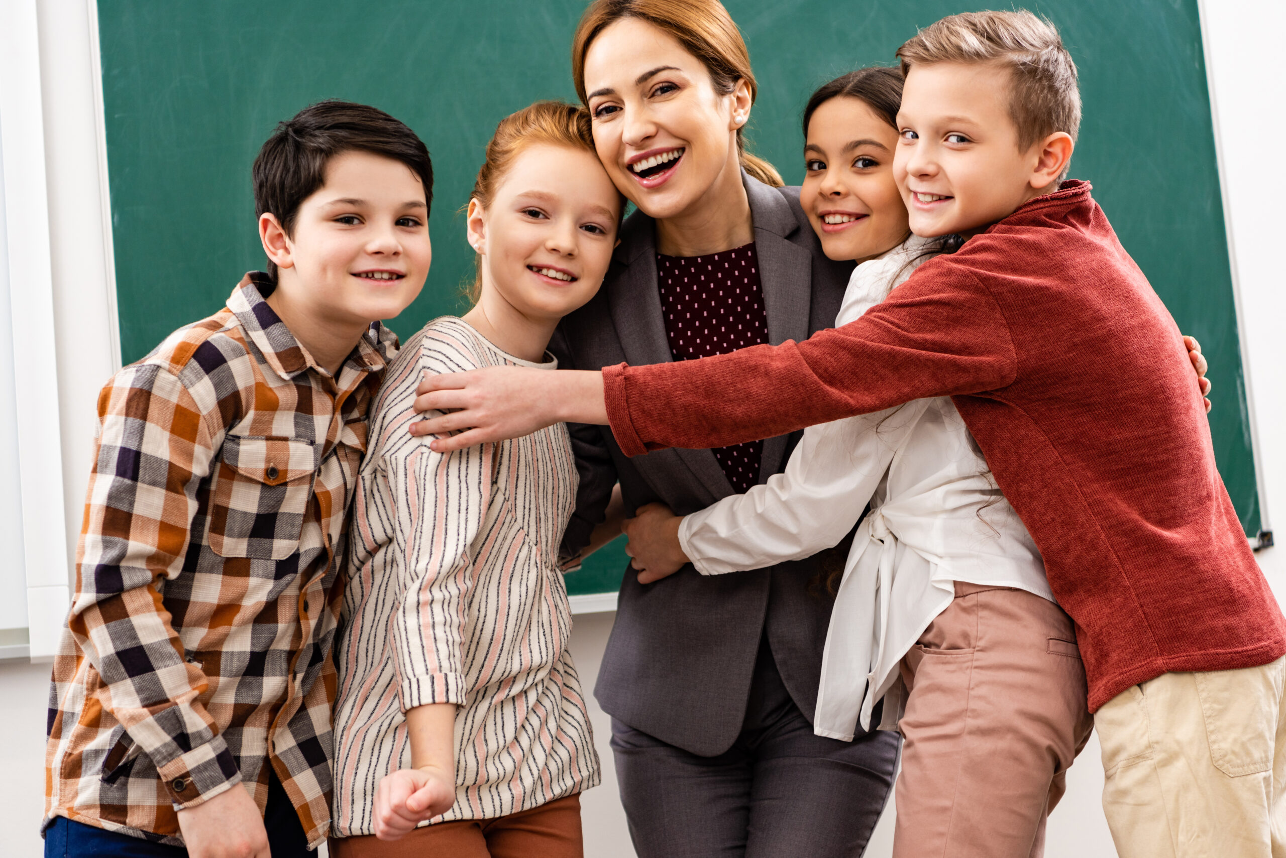 Happy pupils embracing teacher in front of blackboard in classroom